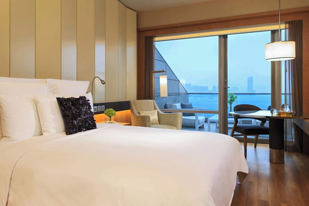 A hotel room with a large bed, a desk, a chair, and a view of the city through large windows.