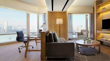 Een moderne hotelkamer met een bank, fauteuil en een ronde salontafel. Er is een televisie aan de muur bevestigd en een groot raam met uitzicht op de stad.