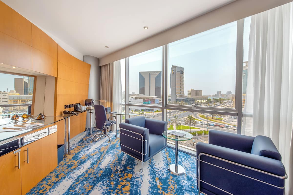 Moderni hotellihuone, jossa on suuri ikkuna, josta avautuu näkymä kaupunkiin, pieni ruokailutila sekä oleskelualue, jossa on kaksi nojatuolia ja pöytä.