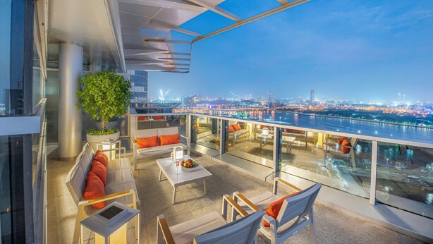 A modern rooftop lounge with a city view, seating areas, and a potted plant.