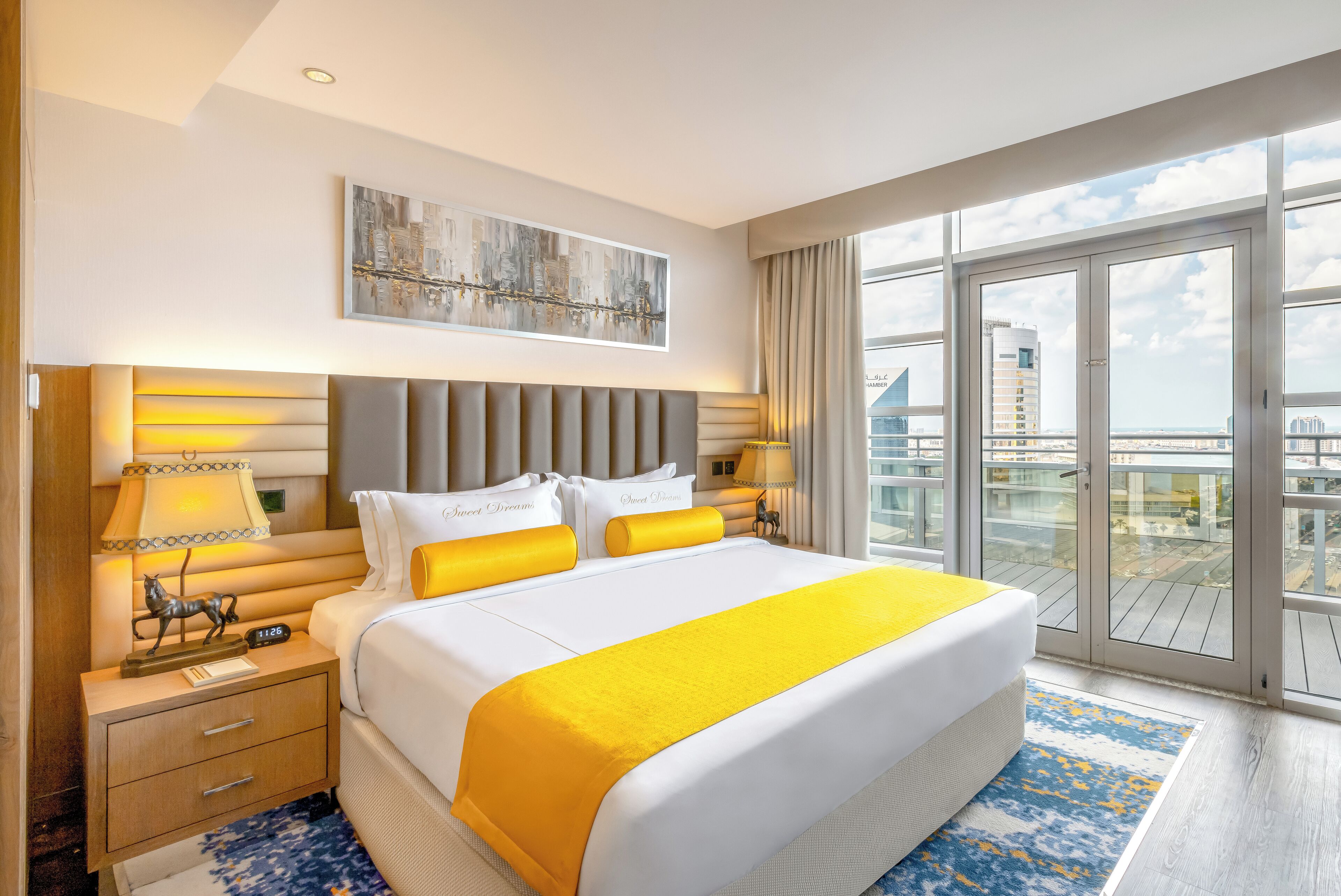 A hotel room with a large bed, a nightstand, a lamp, and a view of the city through large windows.