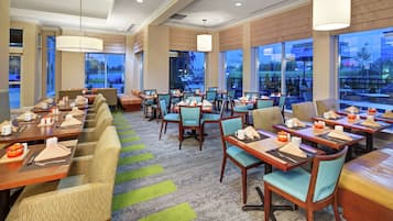 A hotel dining area with multiple tables and chairs, large windows, and a view of the outside.
