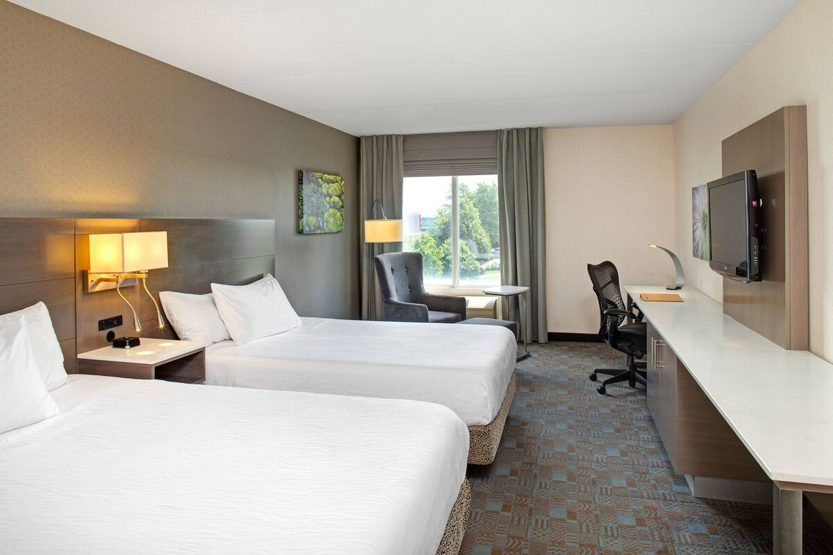 Habitación de hotel con dos camas, un escritorio, una silla, un televisor y una ventana con vistas a zonas verdes.