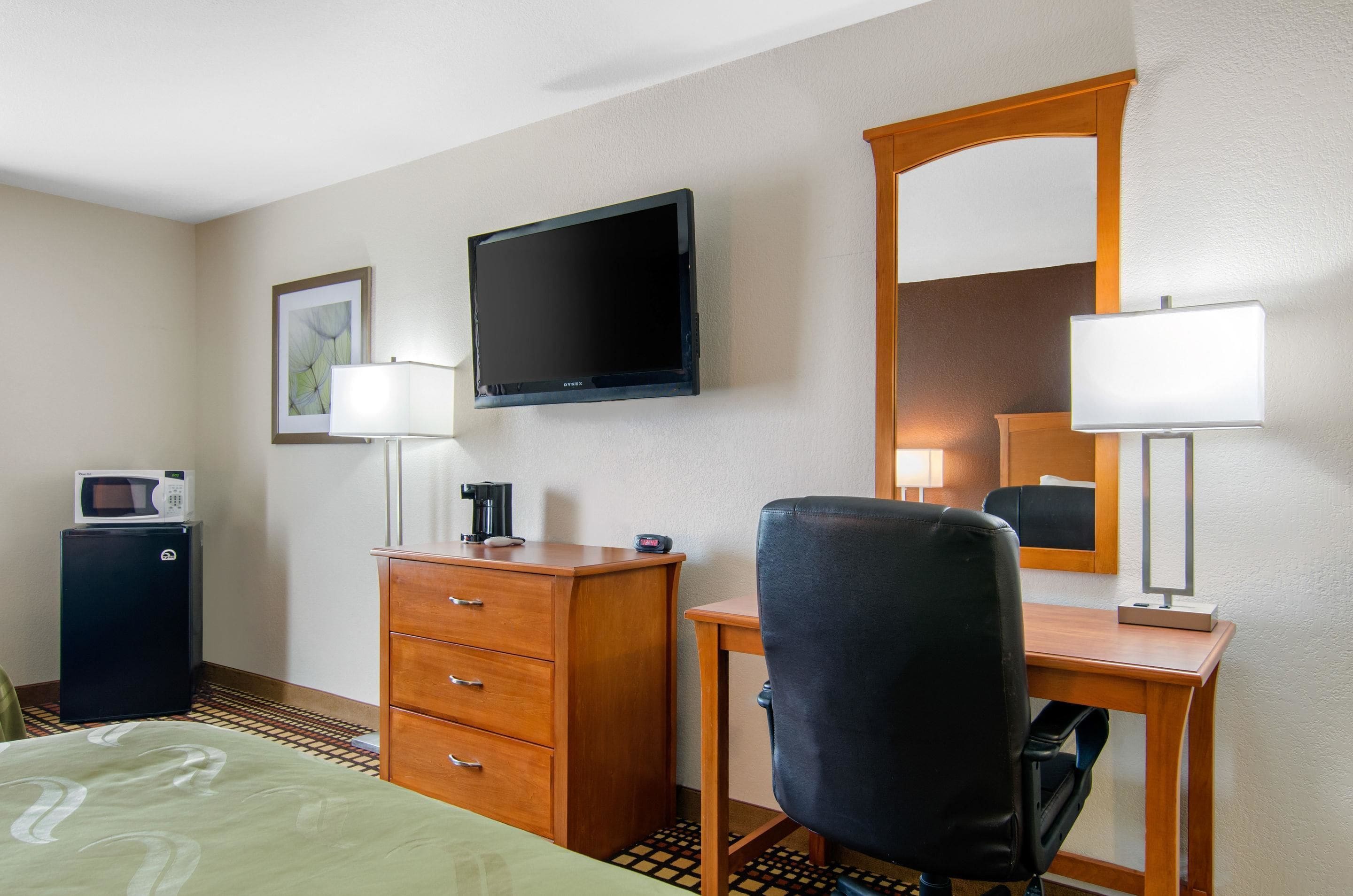 Una habitación de hotel con cama, escritorio y silla, televisión, espejo, cómoda y electrodomésticos como microondas y cafetera.