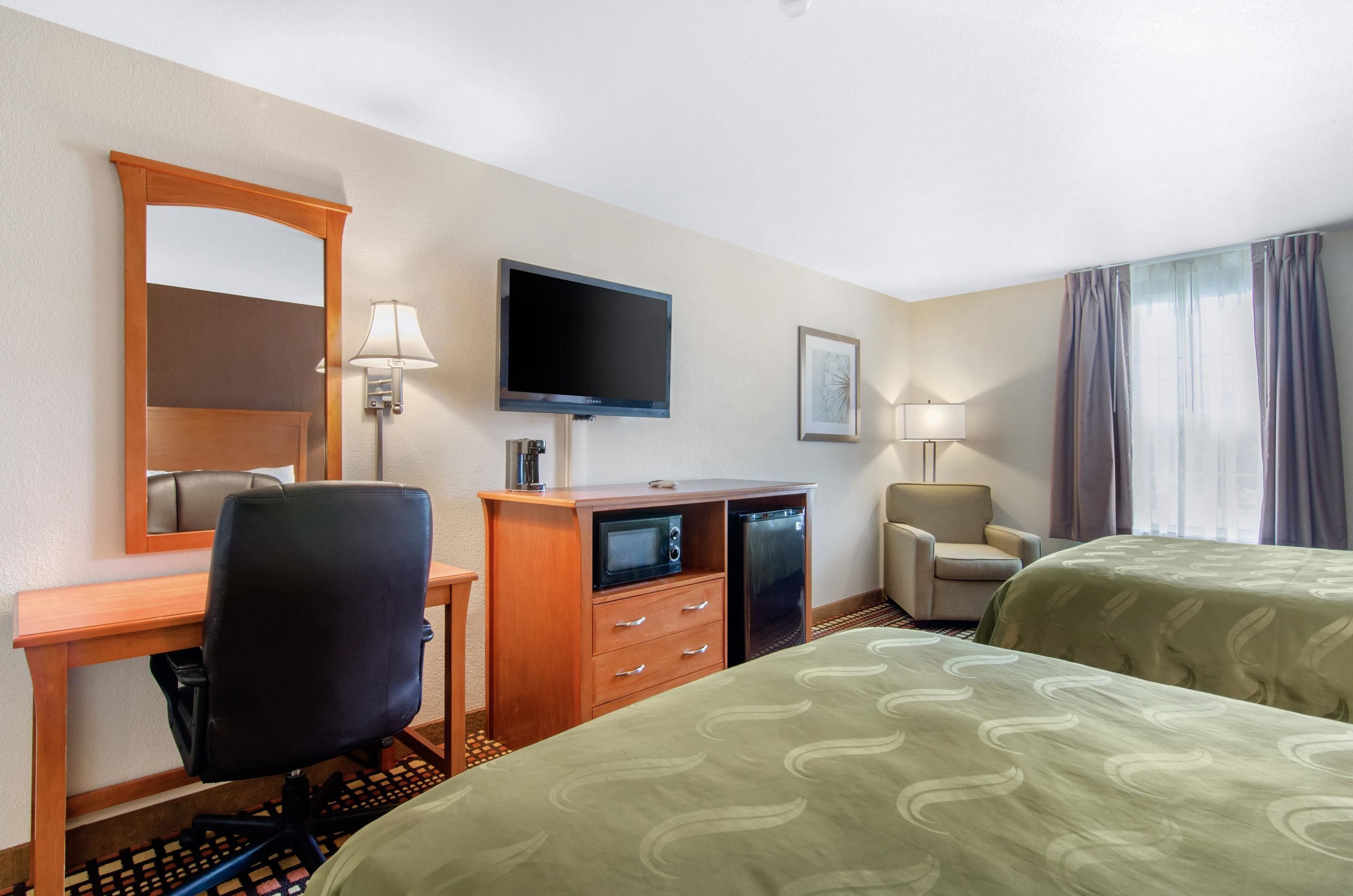 A hotel room with two beds, a desk with a chair, a television, a mirror, and a window with curtains.