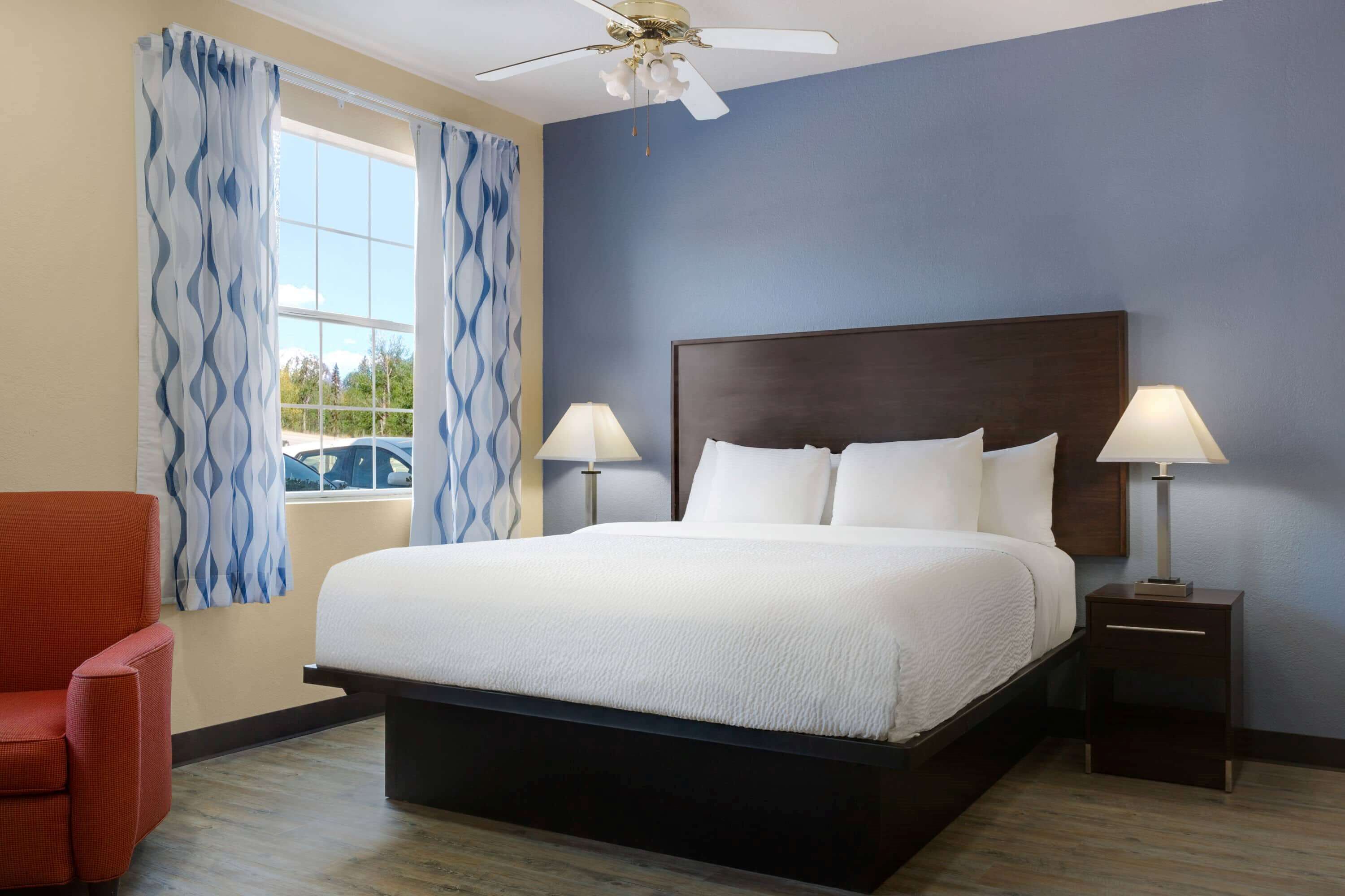 A hotel room with a large bed, two bedside lamps, a ceiling fan, and a view of the outdoors through a window with blue curtains.