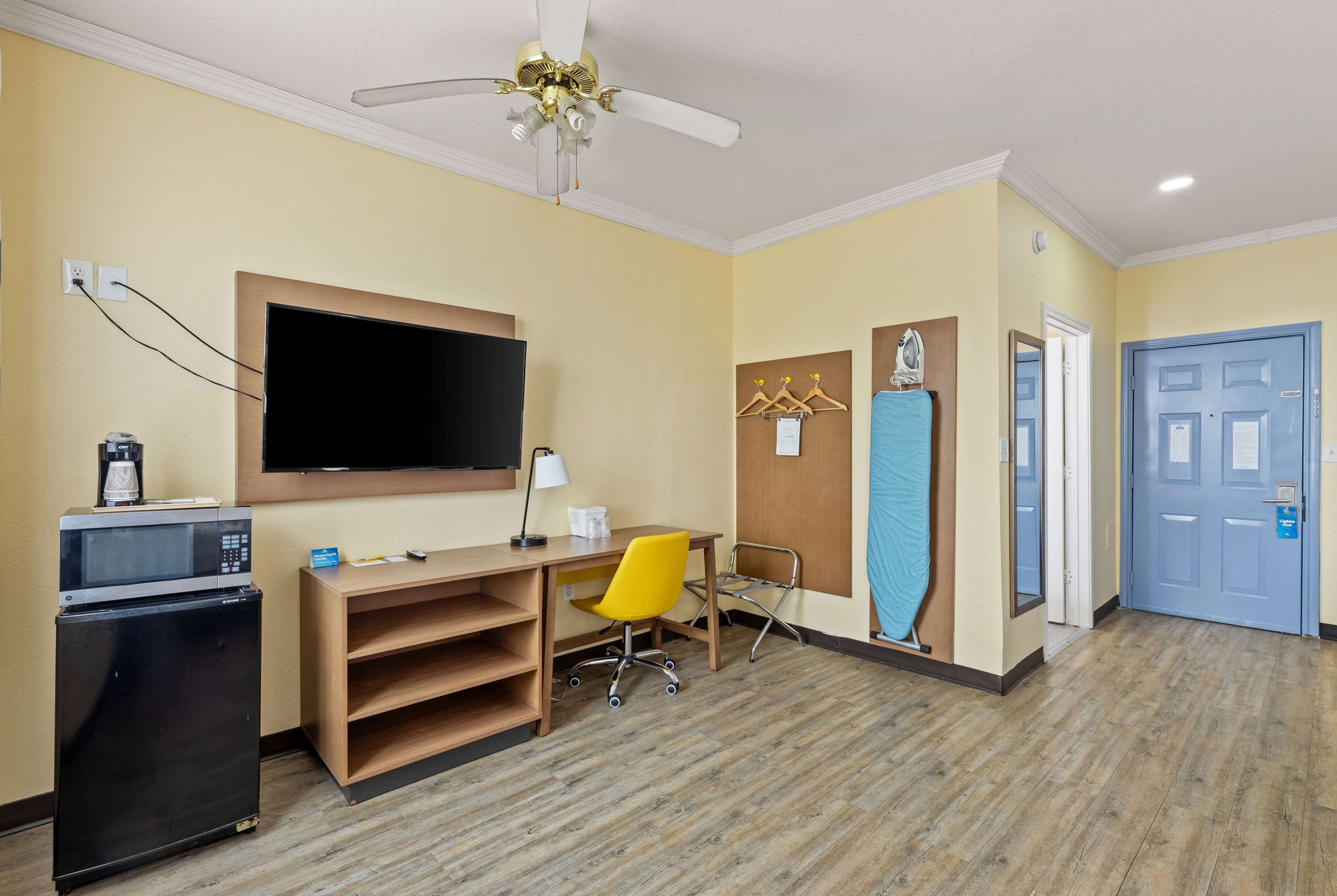 Une chambre d’hôtel équipée d’un bureau, d’une télévision, d’un four à micro-ondes, d’un réfrigérateur, d’une chaise jaune, d’un ventilateur de plafond et d’une porte bleue.