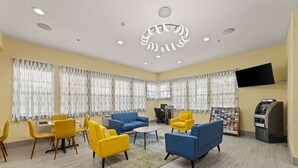 Un hall d’hôtel aux murs jaunes, avec des canapés bleus, des fauteuils jaunes, une table basse ronde et un téléviseur fixé au mur.