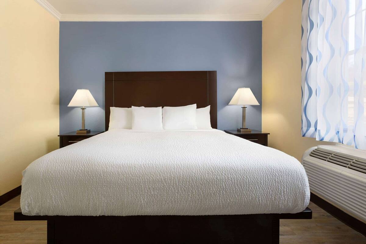 Ein Hotelzimmer mit einem großen Bett, zwei Nachttischen mit Lampen, einem Fenster mit Vorhängen und einer Wand-Klimaanlage.