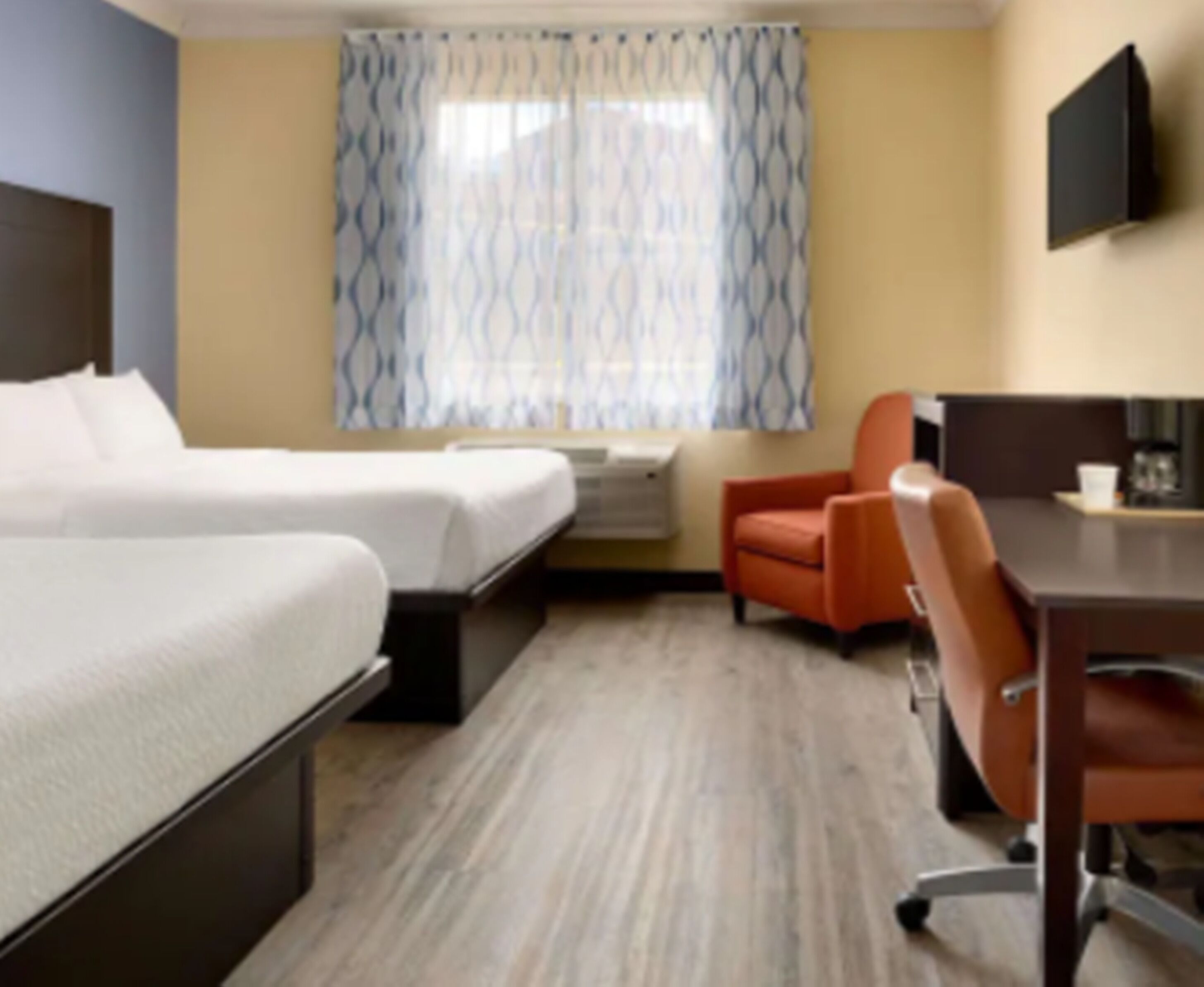 Habitación de hotel con cama, escritorio, una silla naranja y televisor en la pared.