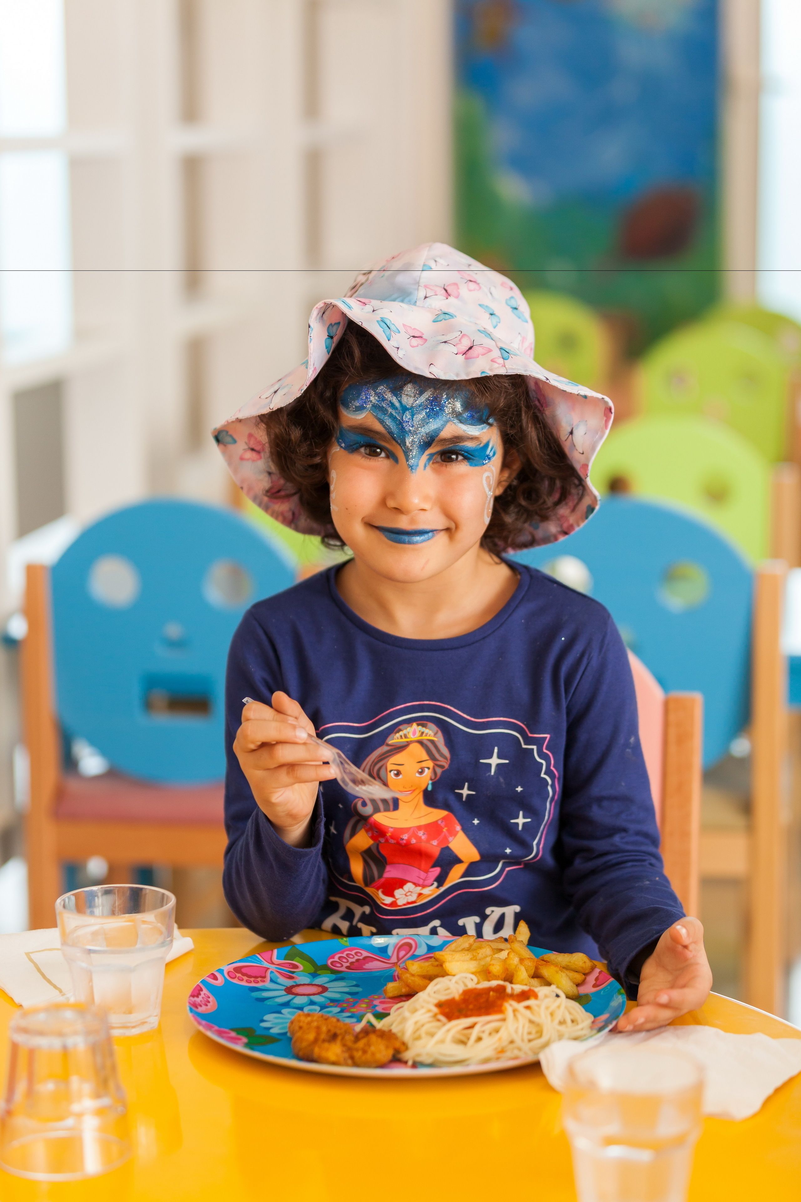A child sitting at a table with a plate of food, a glass of water, and a colorful hat.