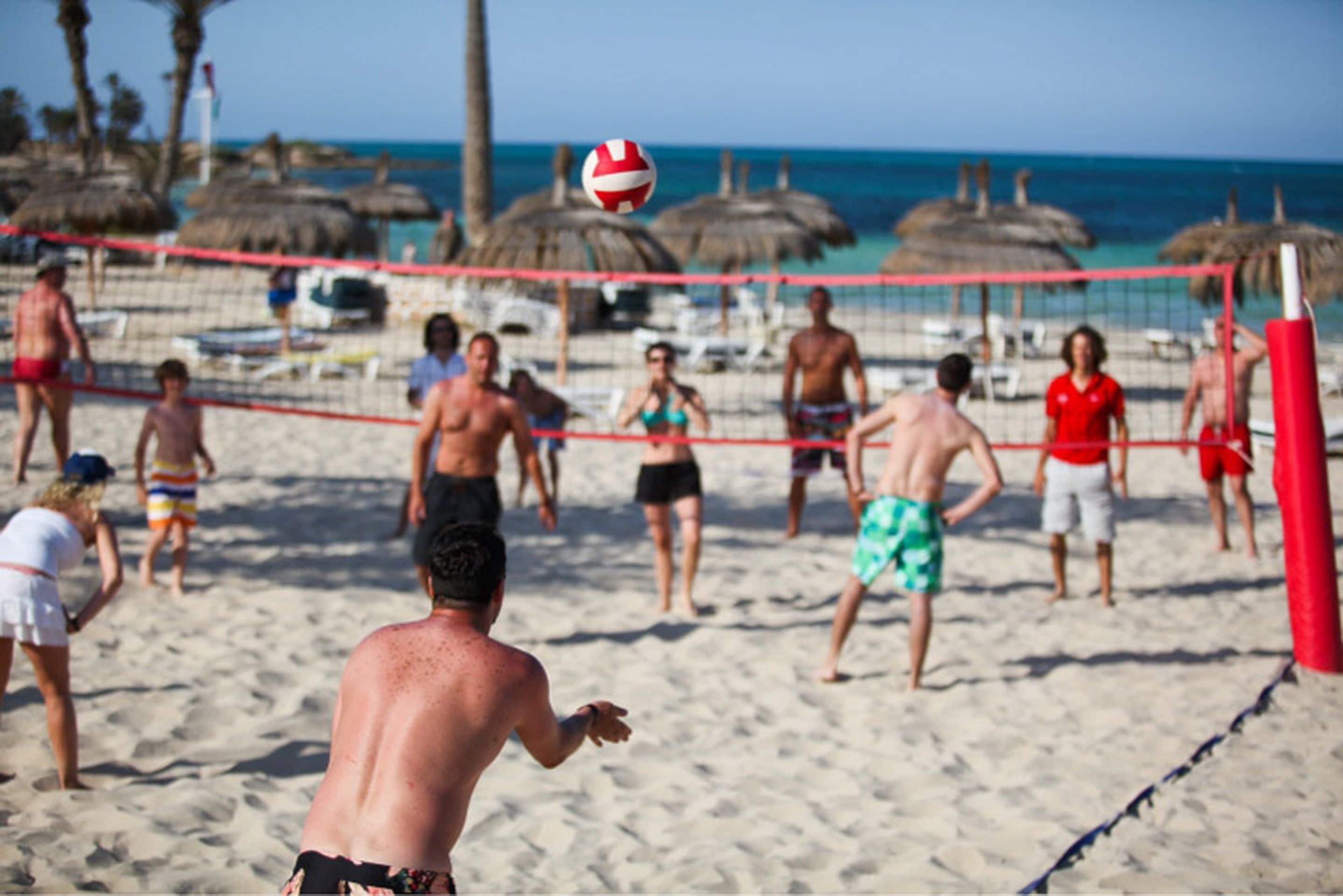 A beach volleyball game with players on a sandy court, a net, and beach umbrellas in the background.