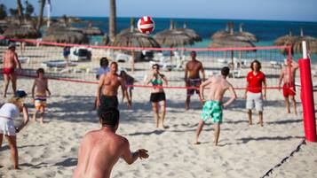 A beach volleyball game with players on a sandy court, a net, and beach umbrellas in the background.