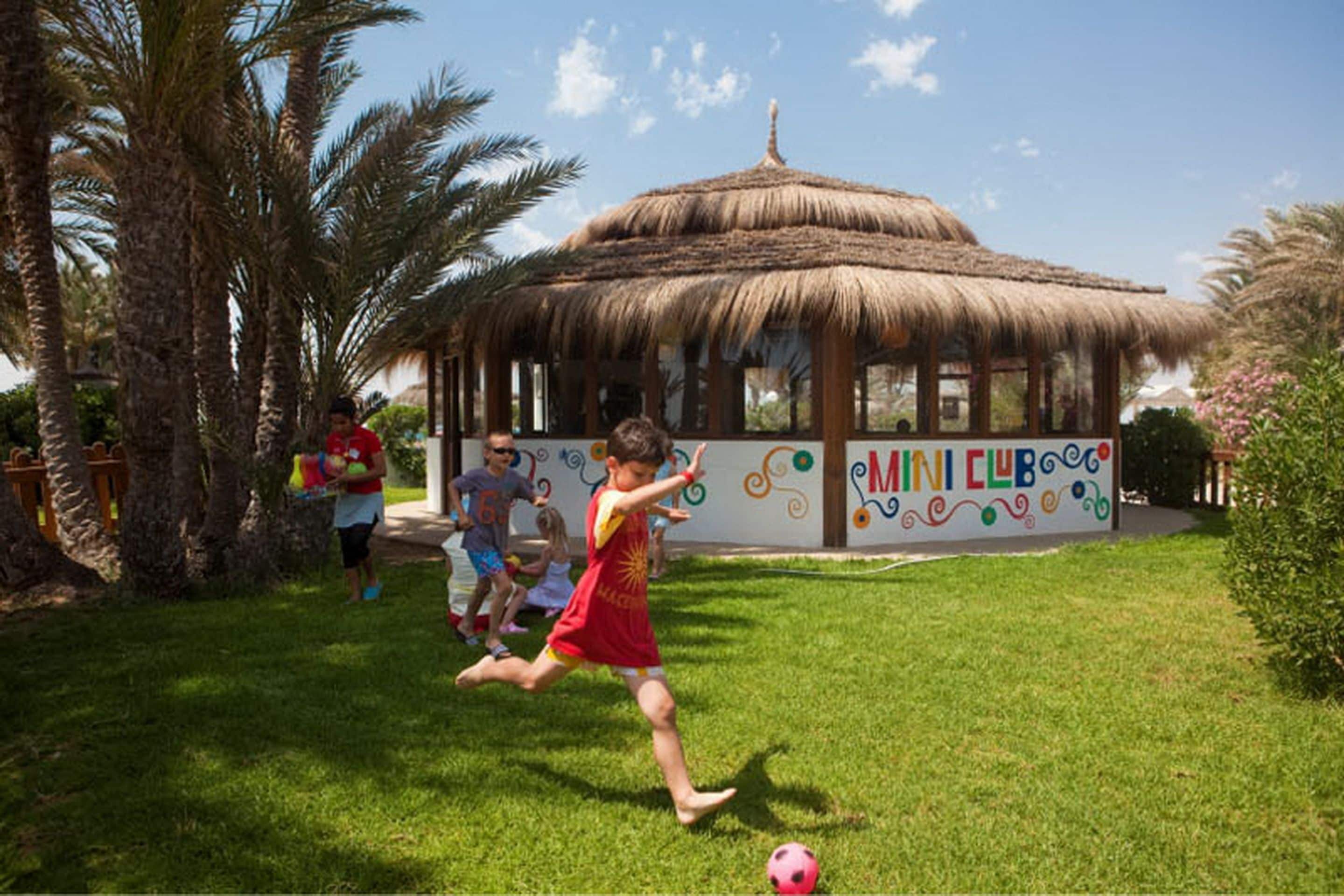A thatched-roof pavilion with a colorful mural labeled "MINI CLUB" in a grassy area with palm trees.