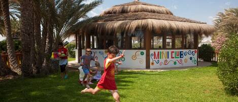 A thatched-roof pavilion with a colorful mural labeled "MINI CLUB" in a grassy area with palm trees.