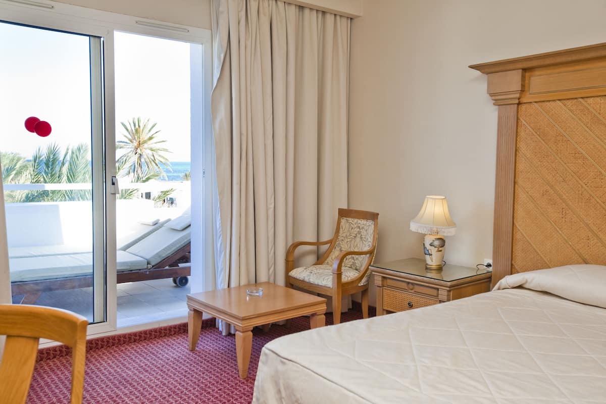 A hotel room with a bed, a chair, a small table, a nightstand with a lamp, and a view of a pool and palm trees.