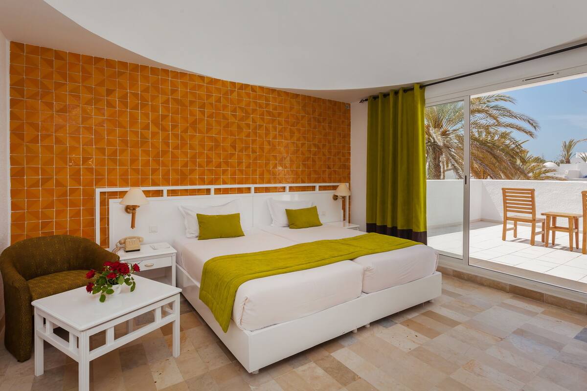 A hotel room with a bed, a small table with a vase of flowers, a chair, and a view of palm trees through a sliding glass door.