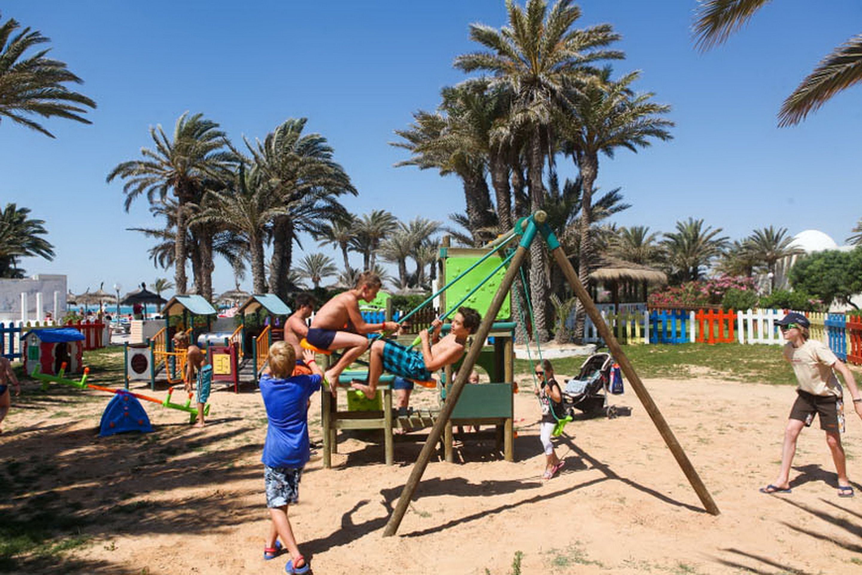 Un parque infantil con columpios y estructuras para escalar, rodeado de palmeras y una zona de arena.