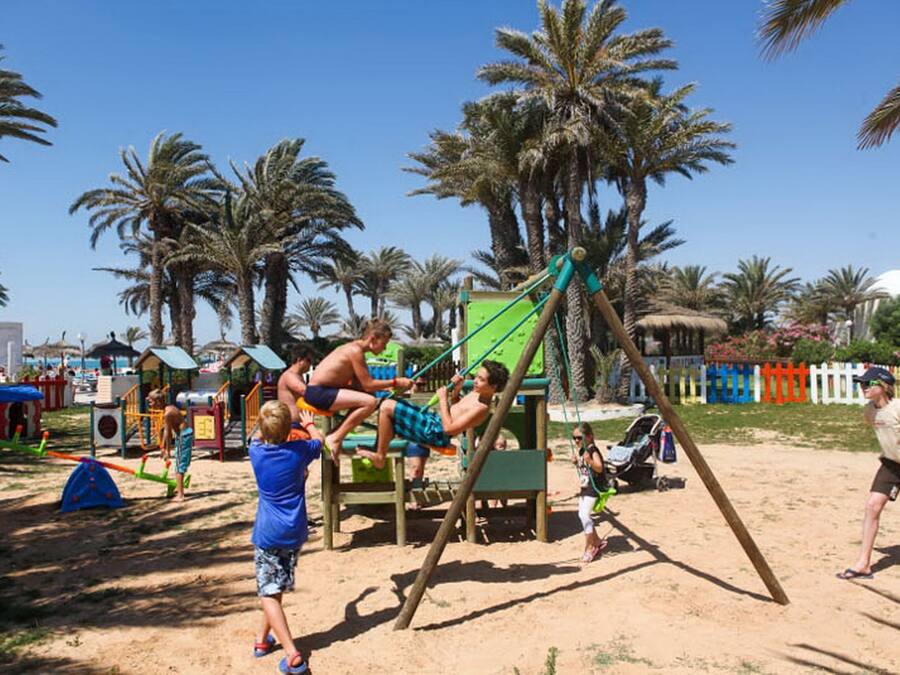 Un parque infantil con columpios y estructuras para escalar, rodeado de palmeras y una zona de arena.