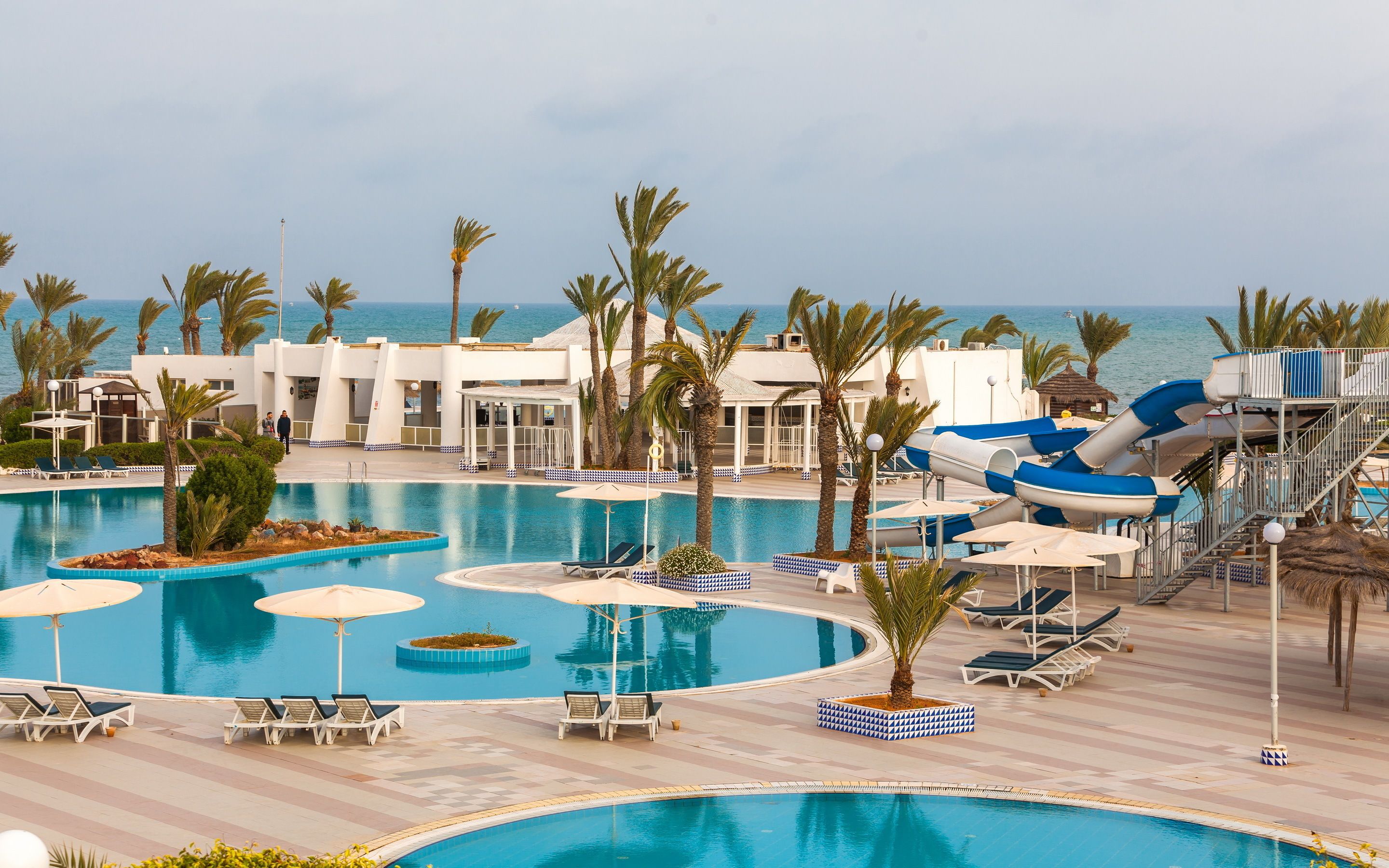 A resort pool area with multiple swimming pools, lounge chairs, palm trees, and a water slide.