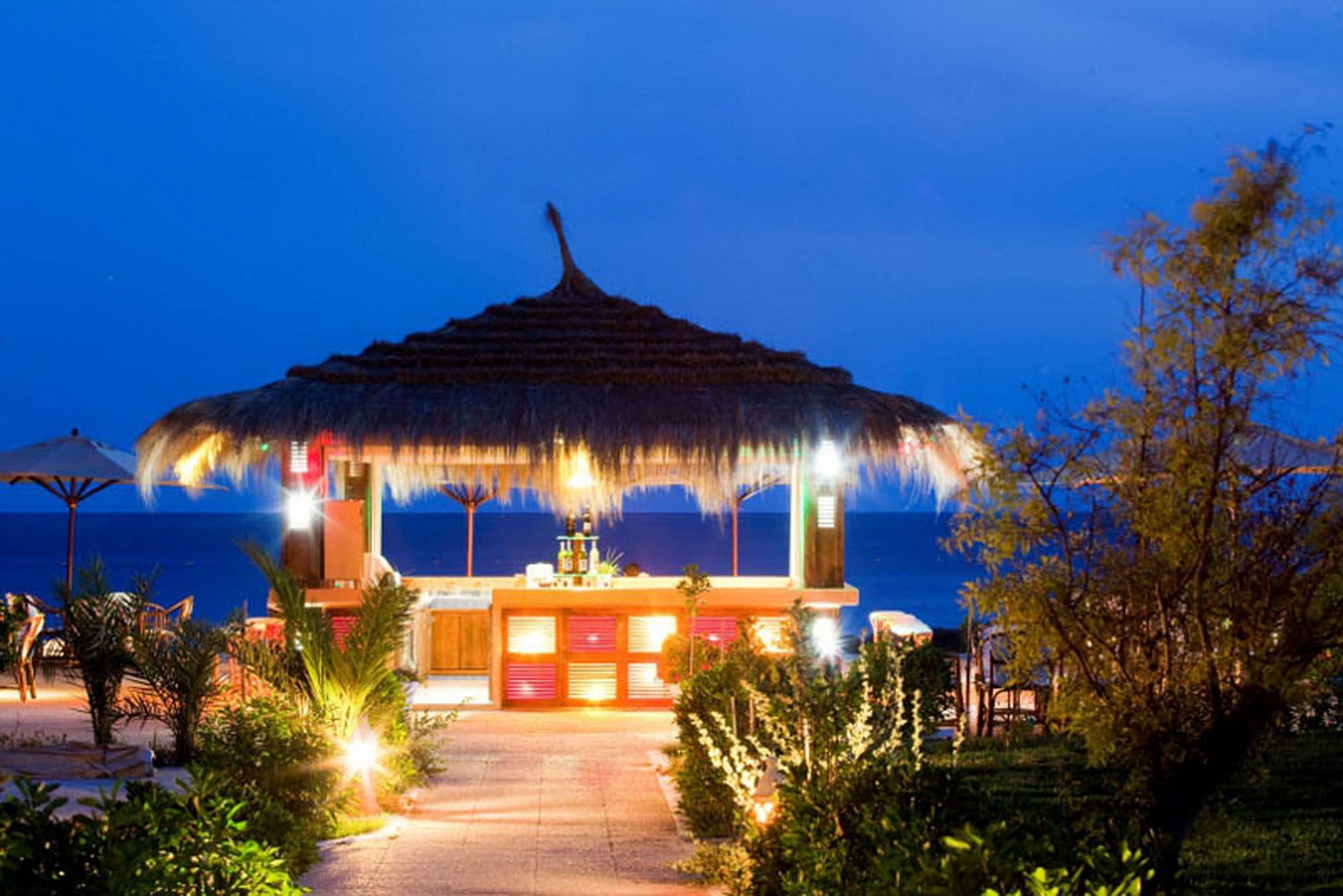 A thatched-roof outdoor bar area with a view of the sea, surrounded by greenery and a paved walkway.