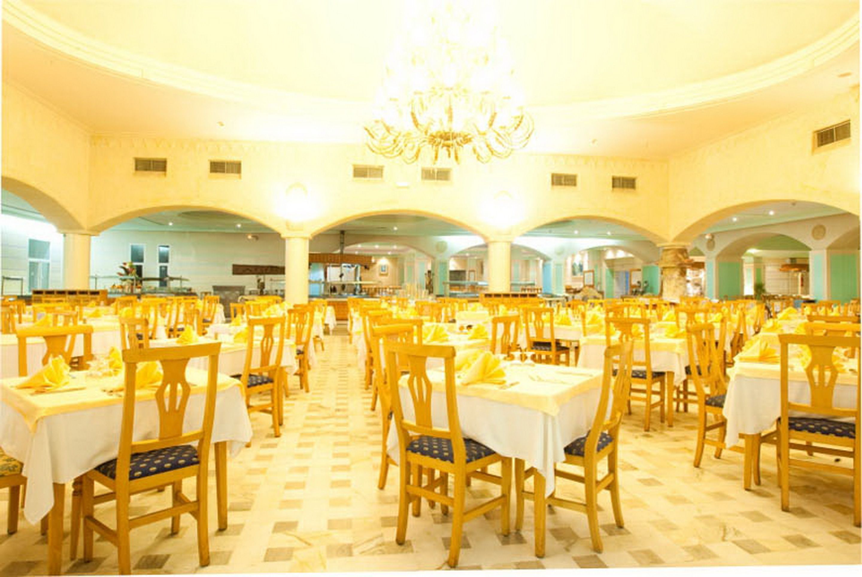 A large dining hall with multiple tables and chairs, a chandelier, and a checkered floor.