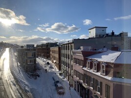 Une rue enneigée bordée d’immeubles, un hôtel bien visible portant le nom de « TELUS », et un ciel dégagé où le soleil brille.