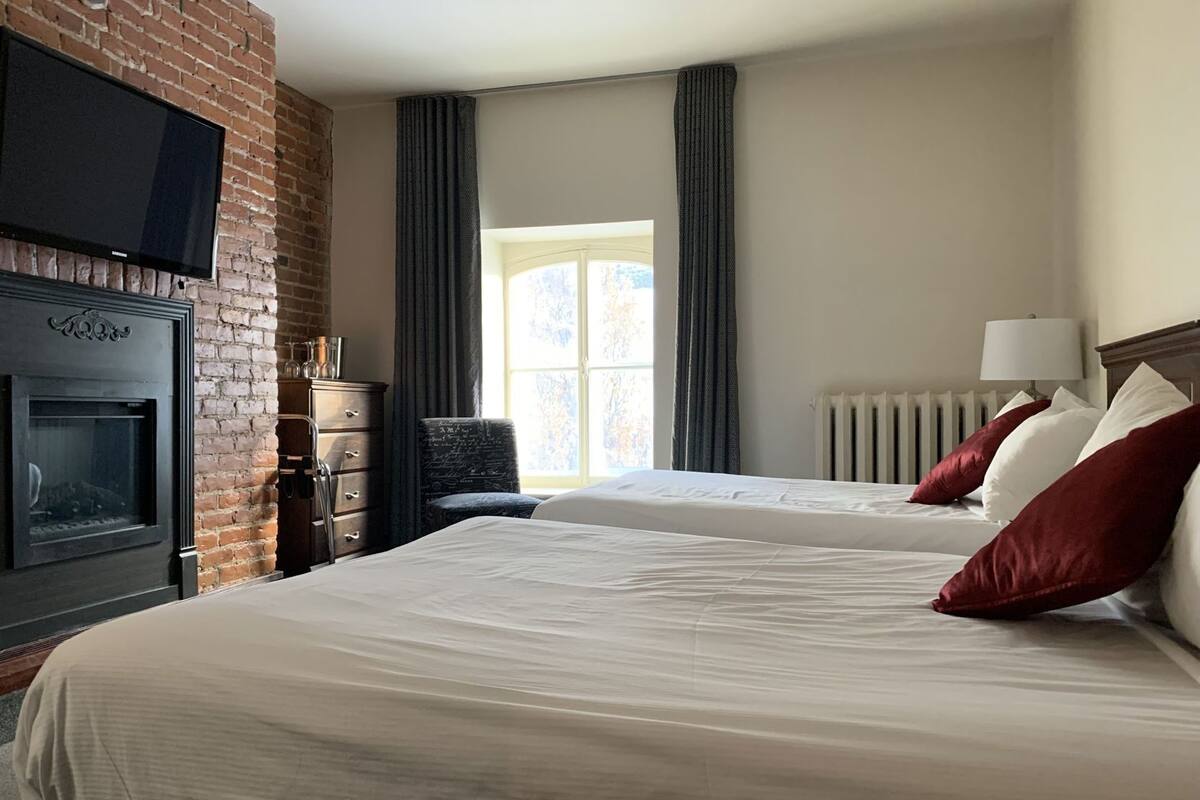 A hotel room with a brick wall, a fireplace, two beds with red pillows, a window, and a TV mounted on the wall.