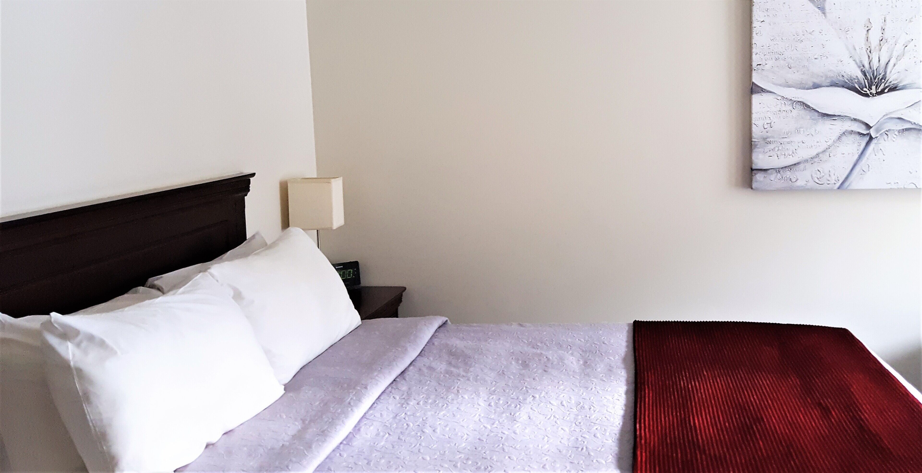 Uma cama bem arrumada com roupa de cama branca e vermelha, uma luminária de cabeceira e uma obra de arte na parede.
