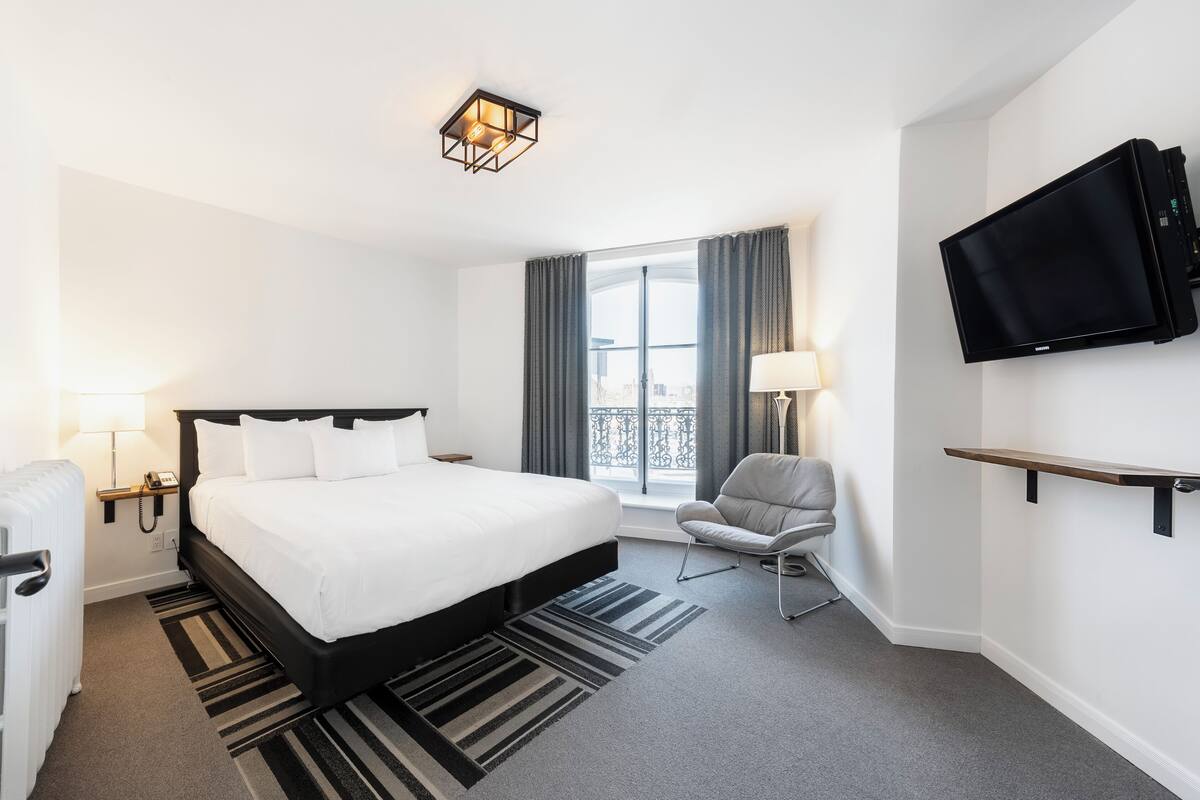 A hotel room with a large bed, a chair, a TV, and a window with curtains.