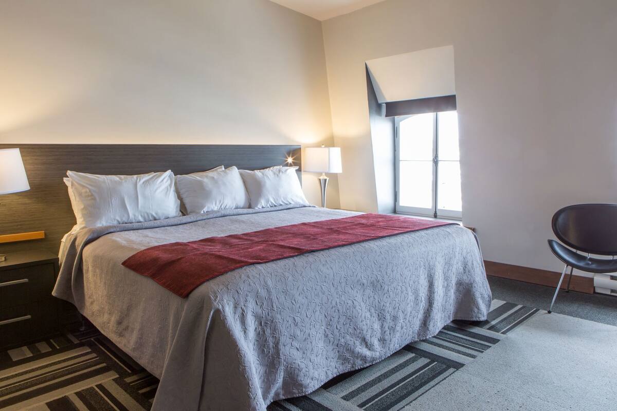 A hotel room with a bed, a desk, a chair, a window, and a lamp.