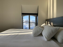 Habitación de hotel con una cama grande, ropa de cama blanca, vistas de un paisaje urbano a través de una ventana y un cabecero moderno.