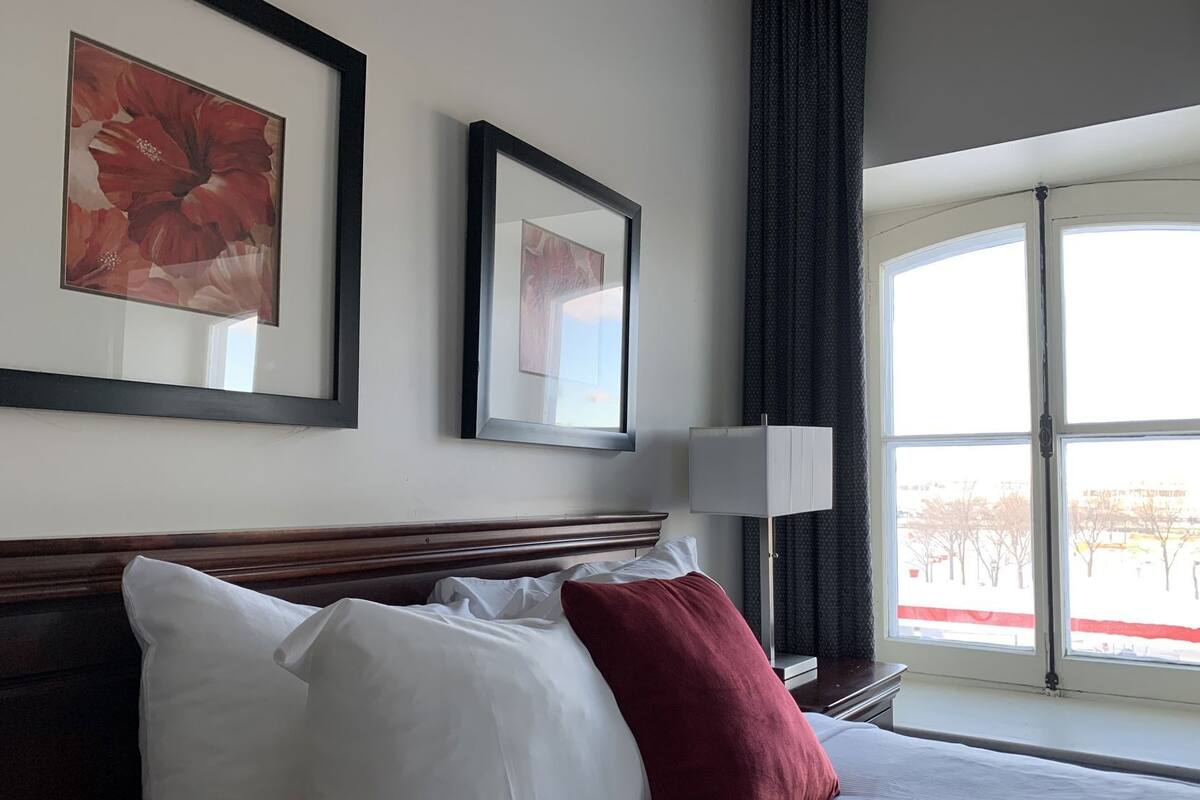 A hotel room with a large bed, two framed artworks on the wall, a window with a view, and a bedside table with a lamp.