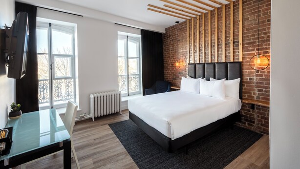 Une chambre d’hôtel moderne dotée d  ’un grand lit, d’un bureau avec une chaise, d’une télévision et d’un mur en briques avec des poutres apparentes.