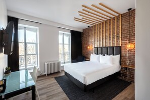 A modern hotel room with a large bed, a desk with a chair, a TV, and a brick wall with exposed beams. - Hôtel Le Saint-Paul (Québec City)