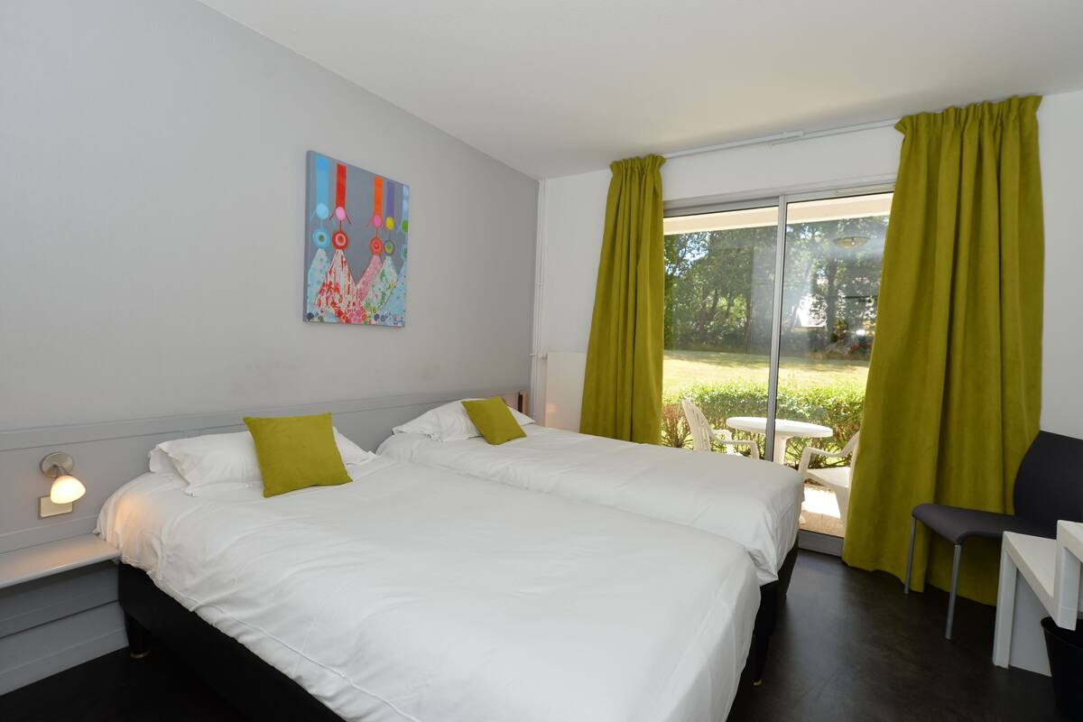 Habitación de hotel con dos camas, un escritorio y una silla. Cuenta con una ventana con cortinas, un cuadro en la pared y una pequeña mesa junto a la ventana.