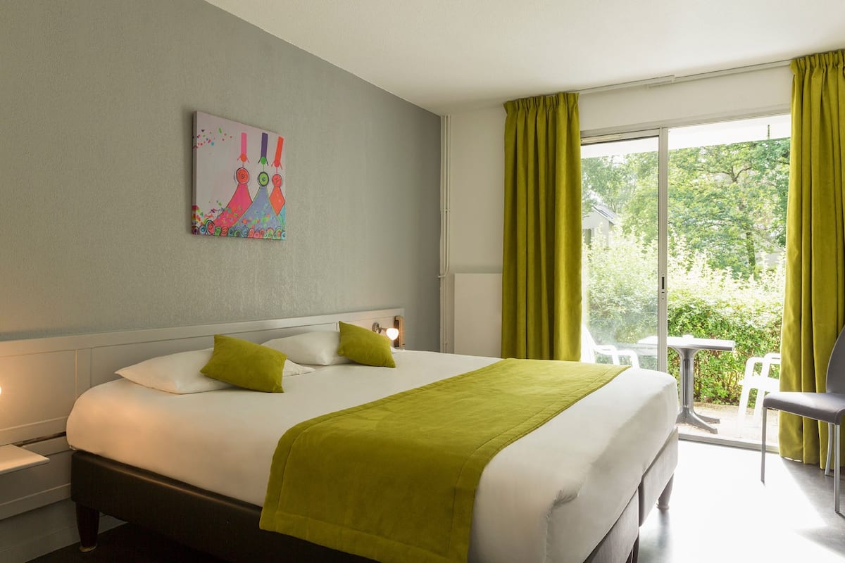 Habitación de hotel con una cama grande, un cuadro en la pared, una ventana con cortinas y vistas a la vegetación.