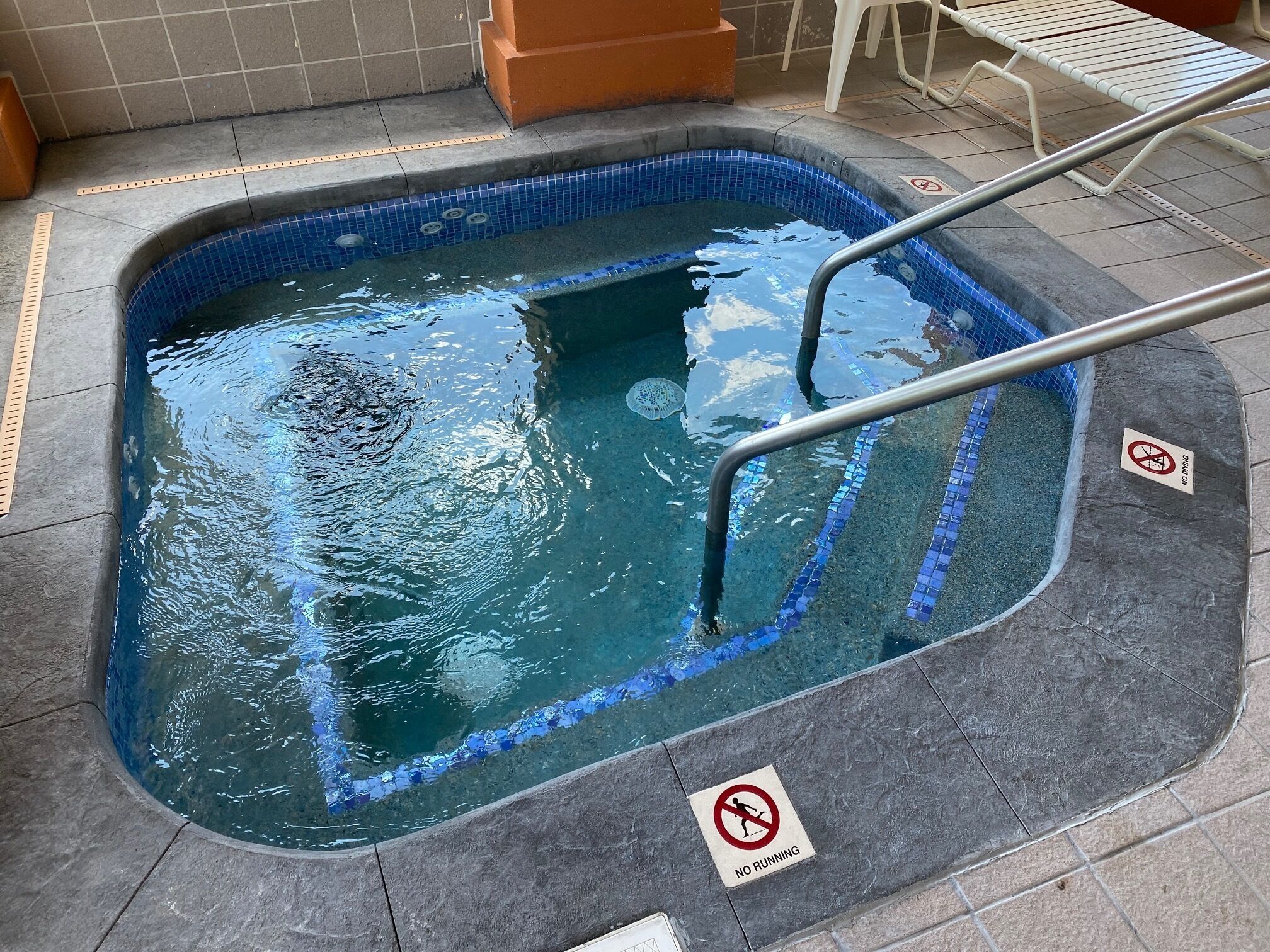 A swimming pool with blue mosaic tiles, metal handrails, and 'NO RUNNING' and 'NO DINING' signs.