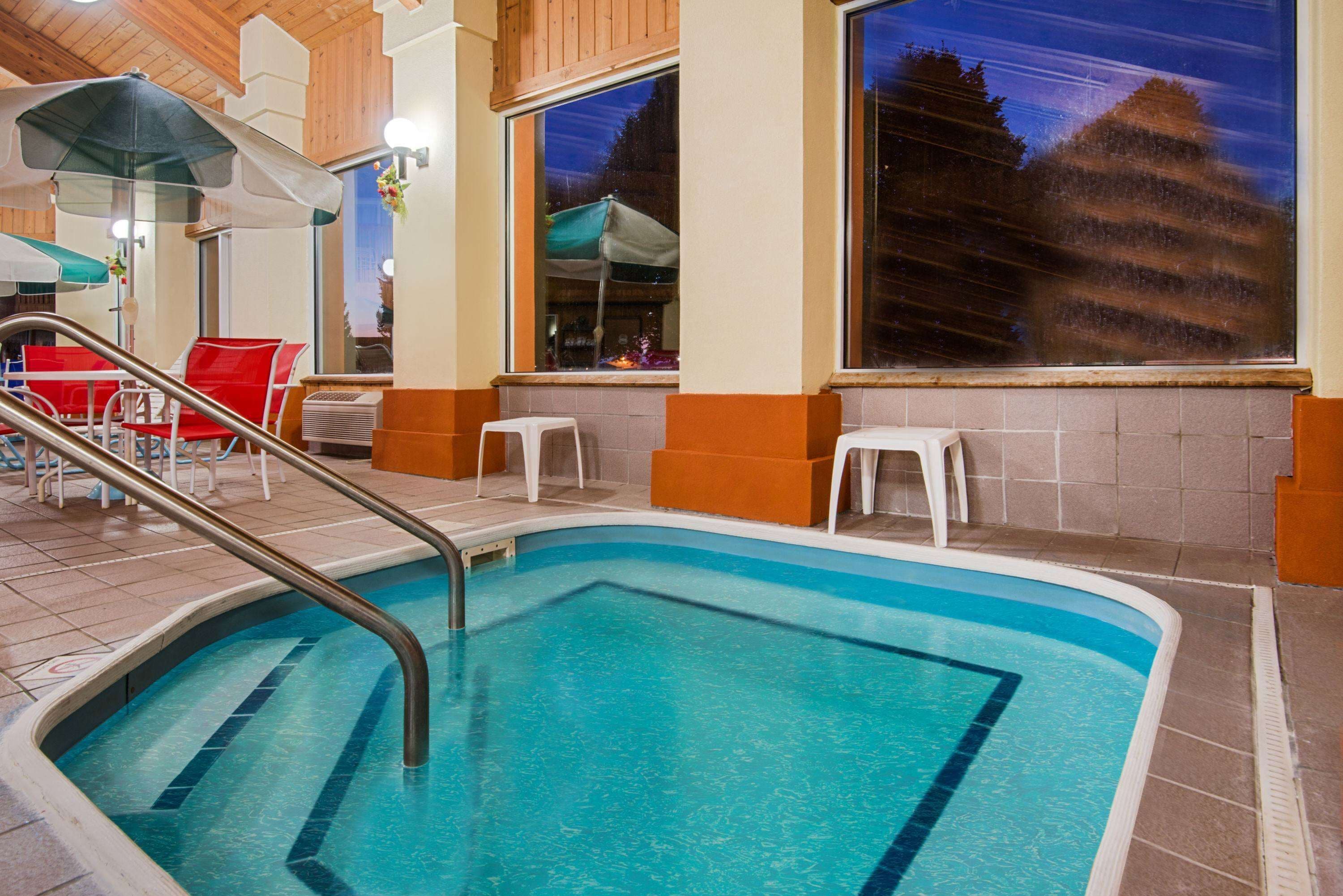A swimming pool with a metal ladder, red chairs, and a view of trees through the windows.