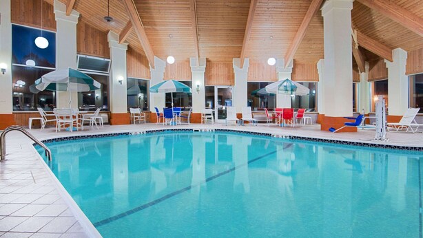 Una piscina interior con agua cristalina, rodeada de sillas de descanso blancas y sombrillas, bajo un techo de madera con grandes ventanales.