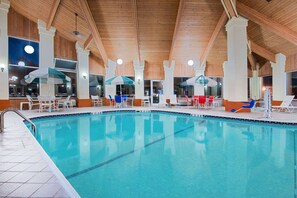 Una piscina interior con agua cristalina, rodeada de sillas de descanso blancas y sombrillas, bajo un techo de madera con grandes ventanales.