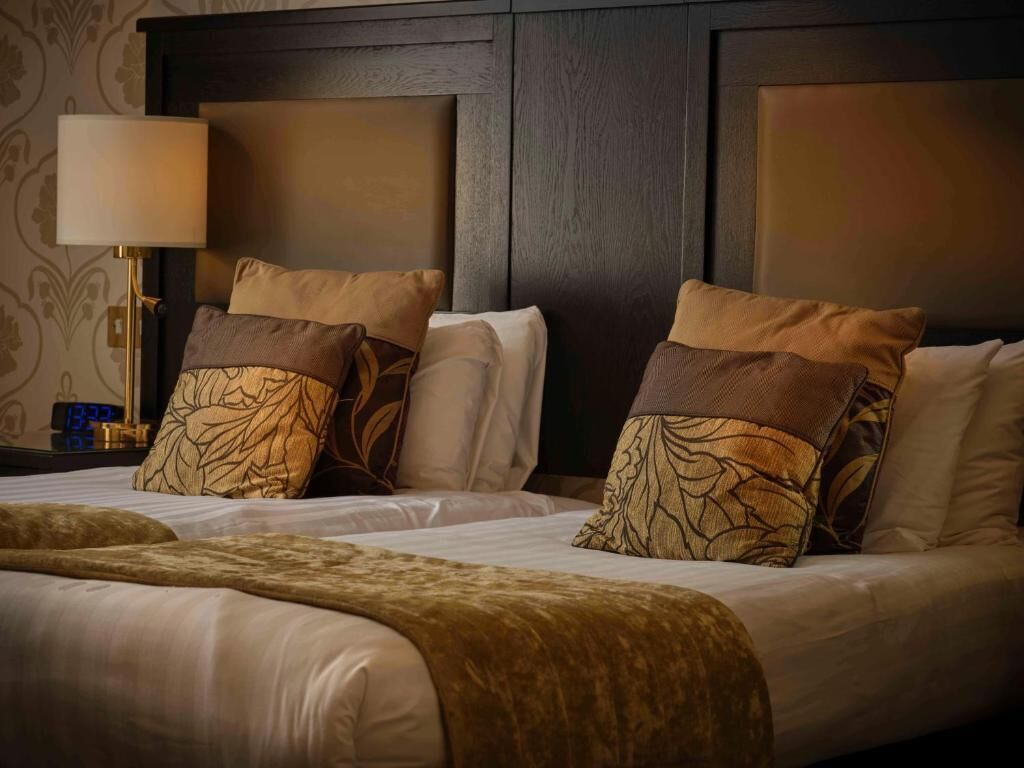 A hotel room with a large bed, two pillows with leaf patterns, a bedside lamp, and a clock on the nightstand.