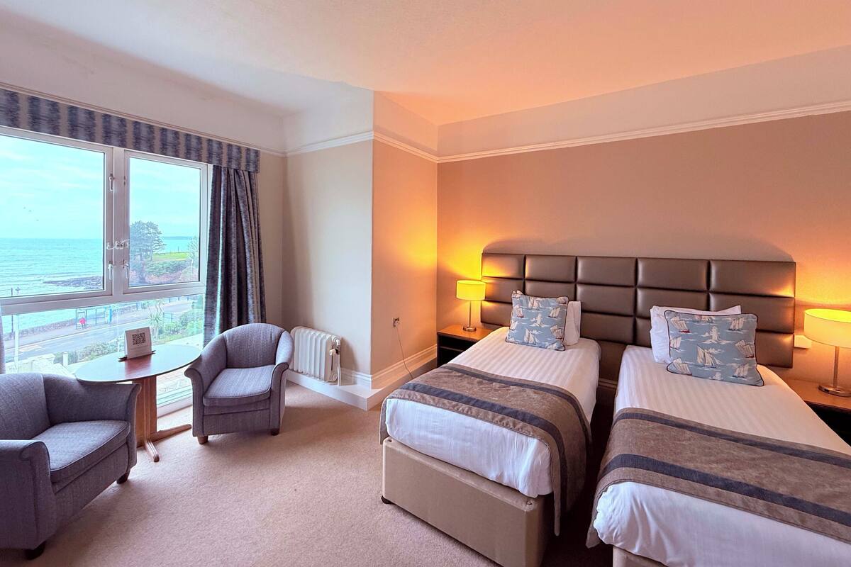 Ein Hotelzimmer mit zwei Betten, einem Sessel, einem Tisch und Blick aufs Meer.