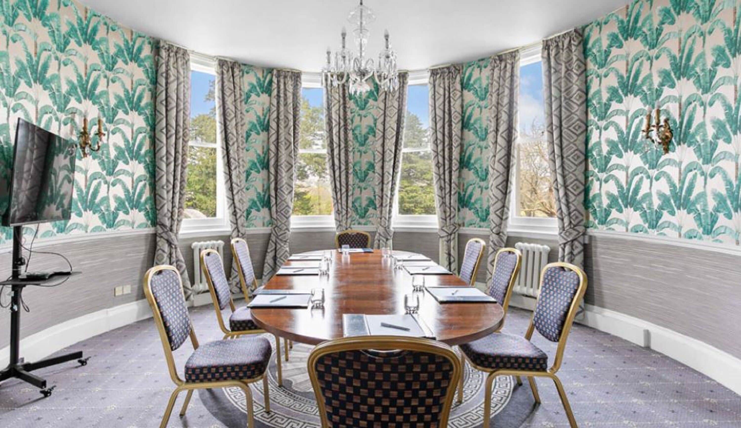 Un salón de conferencias con una mesa redonda, ocho sillas, una araña de cristal y ventanas amplias con cortinas.