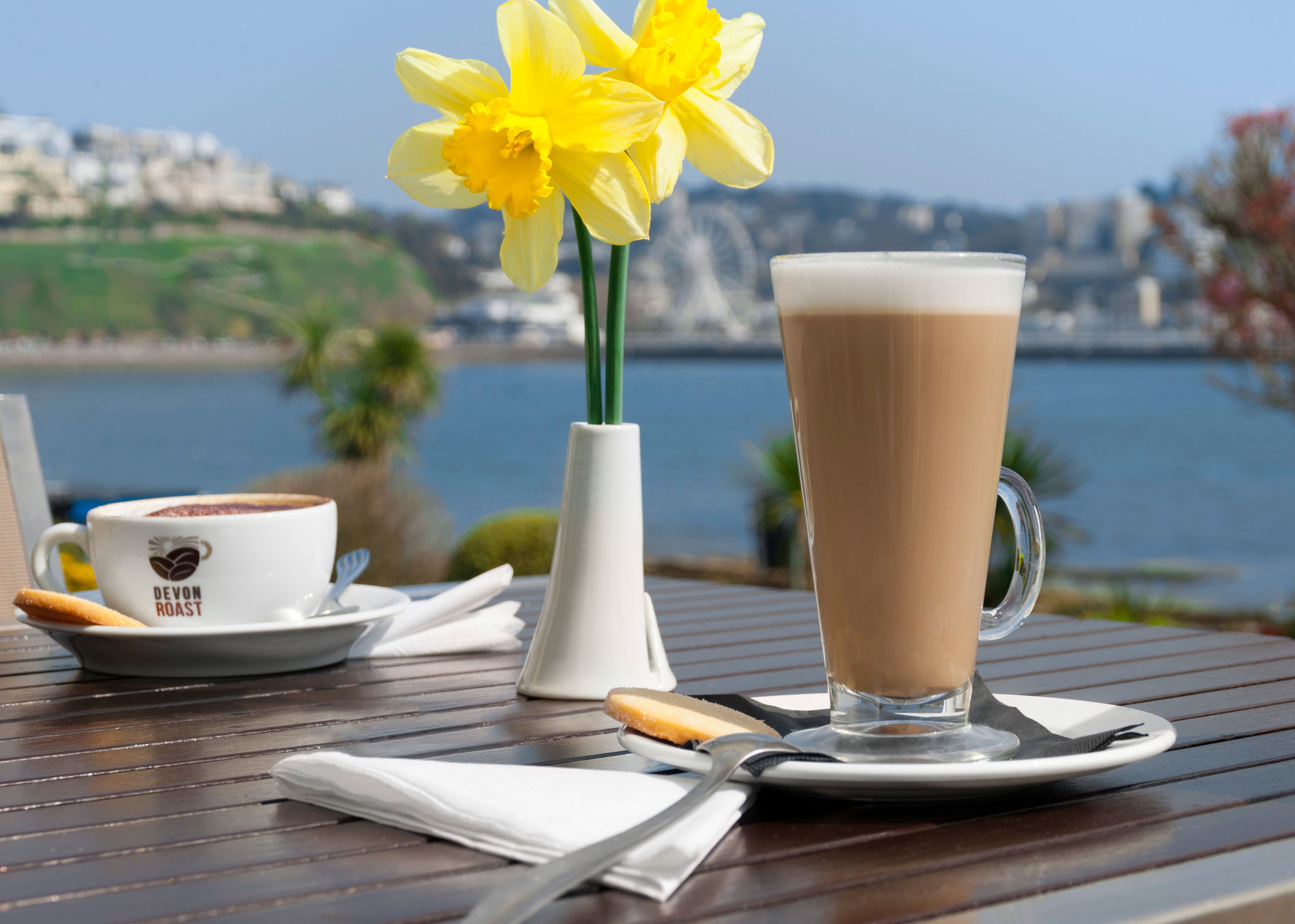 A table with a coffee cup, a tall coffee glass, and a vase with yellow flowers.