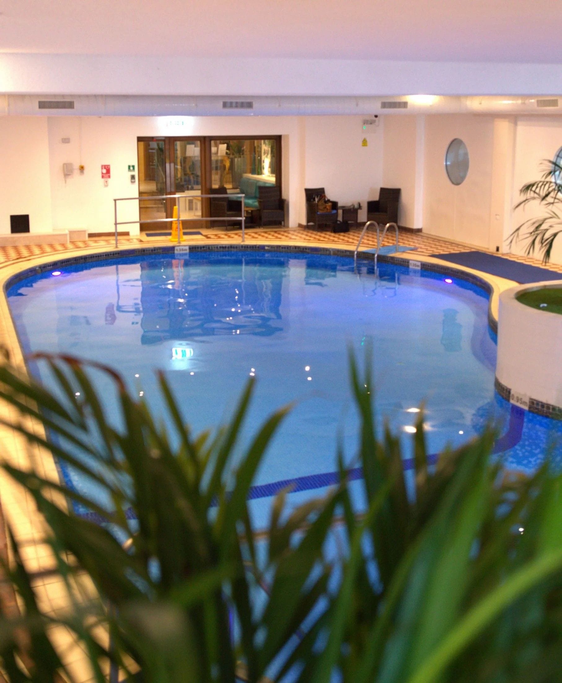 An indoor swimming pool with a clear water surface, surrounded by a tiled deck and a few lounge chairs.