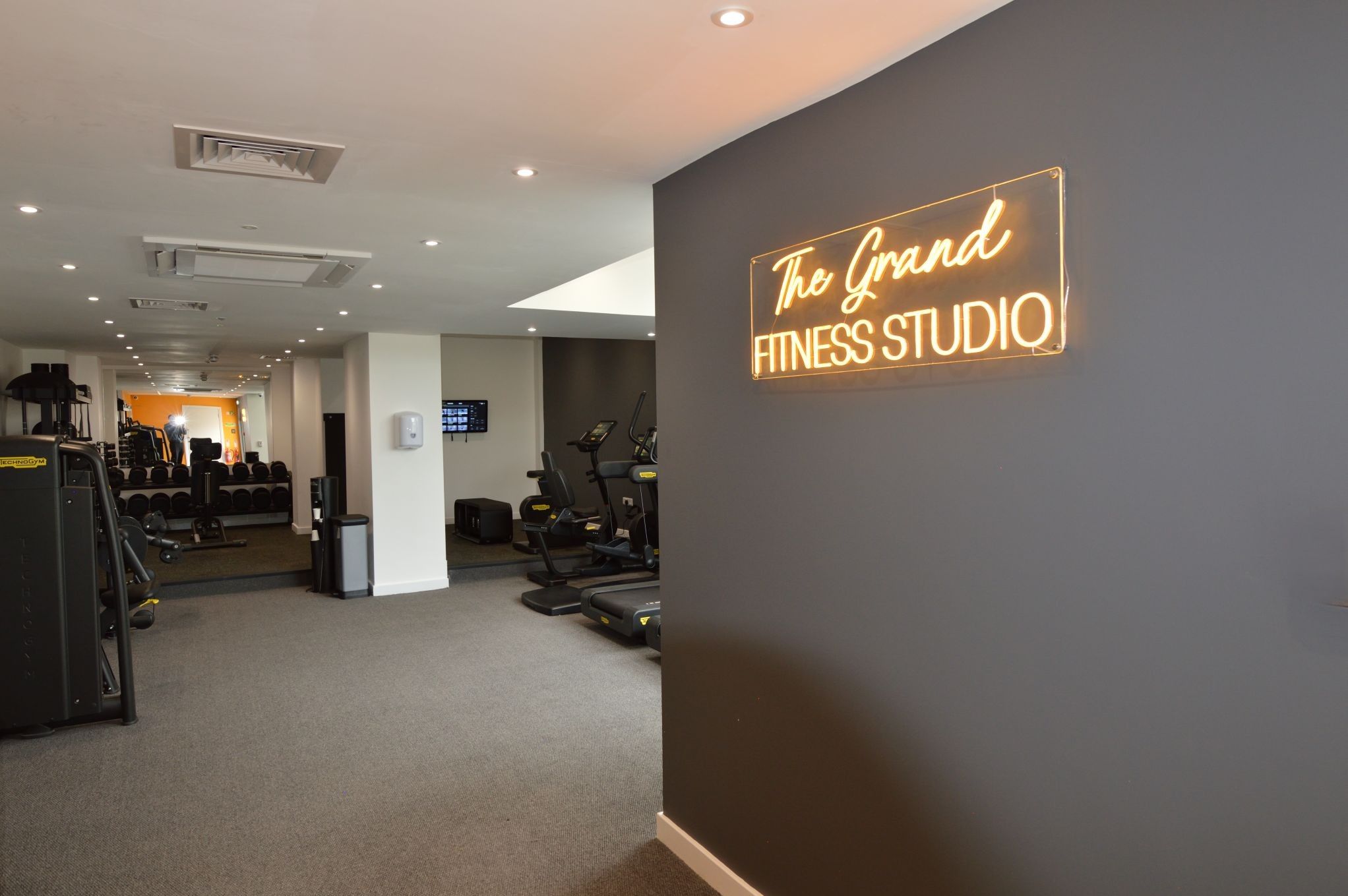 A fitness studio with exercise equipment, a digital display, and a neon sign on the wall.