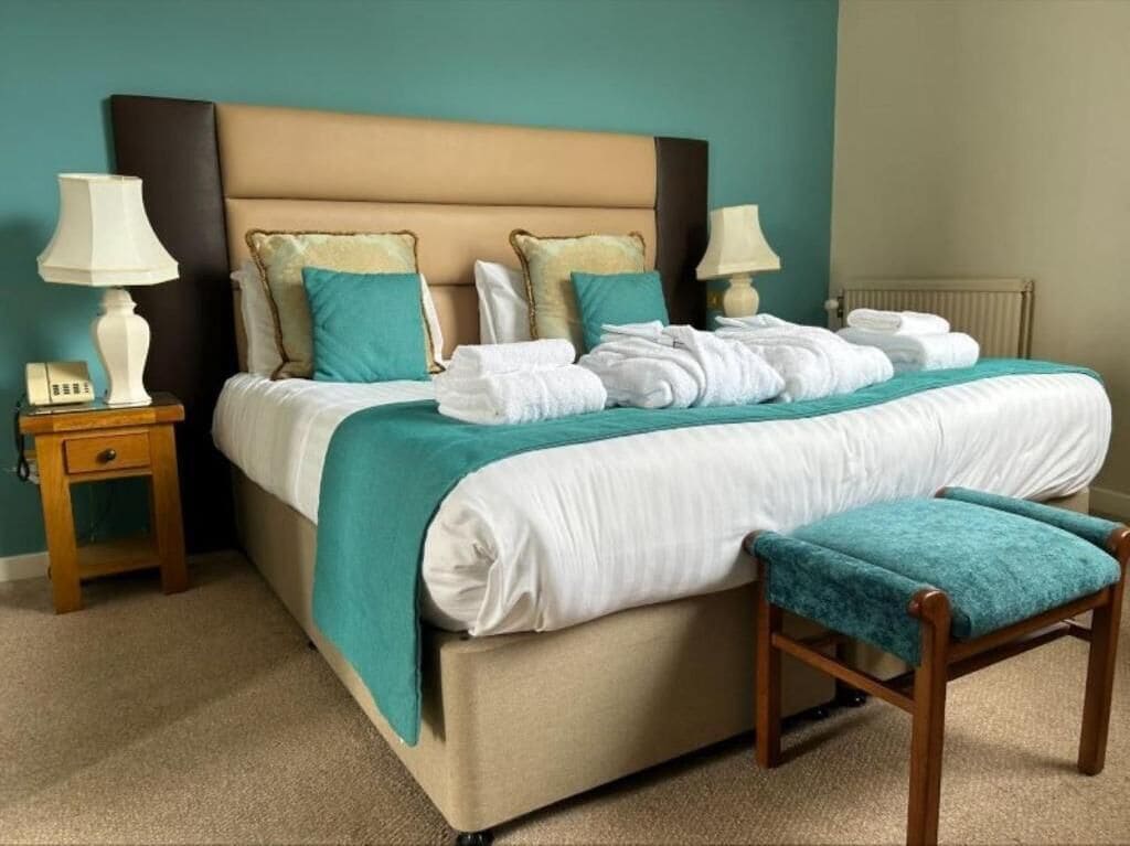 A neatly made bed with a teal bedspread, two bedside lamps, a wooden nightstand, and a matching bench.