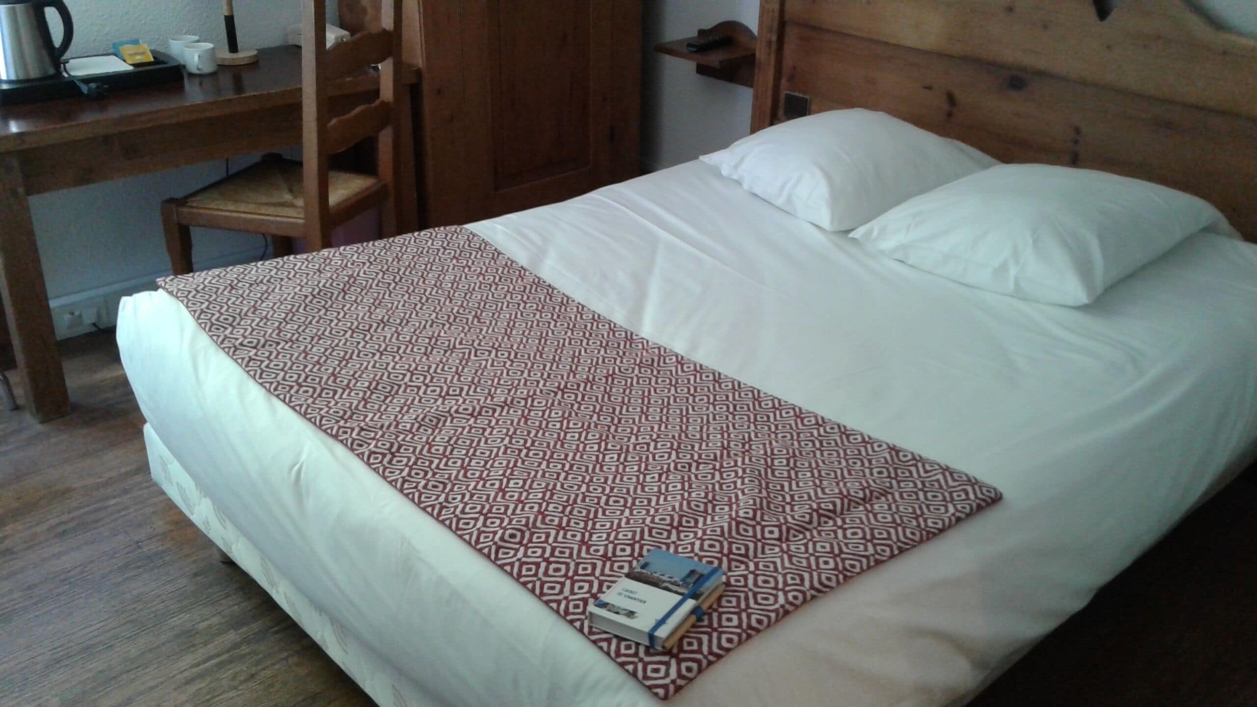 Uma cama com um cobre-cama estampado, uma escrivaninha de madeira com uma chaleira, chávenas e um livro.