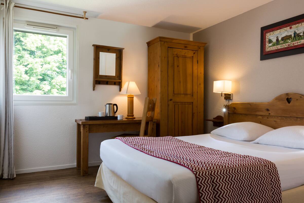 A hotel room with a bed, a wooden wardrobe, a desk with a lamp, and a window with curtains.